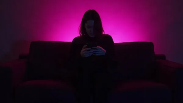 Silueta de espaldas en un sofá sosteniendo un teléfono, en una habitación oscura con resplandor magenta (ilustración del artículo)